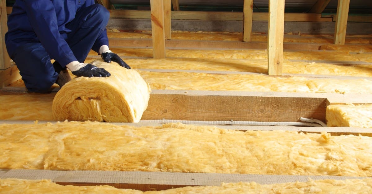 A person wearing a blue jumpsuit is laying insulation in a home attic. The insulation is mustard yellow.