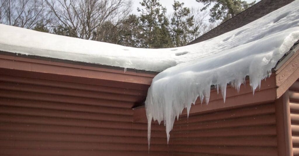 A brown house with ribbed walls holds an ice dam on the edge of its roof. Icicles are seen hanging from the mound of snow.