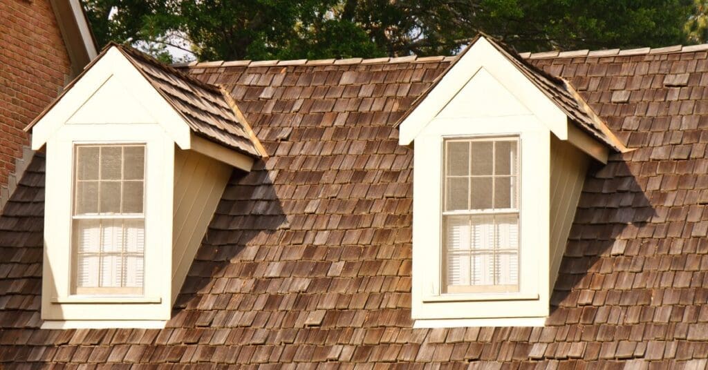 A house has rows of wooden shingles on its roof. Two windows with shades are visible as the sun shines brightly.