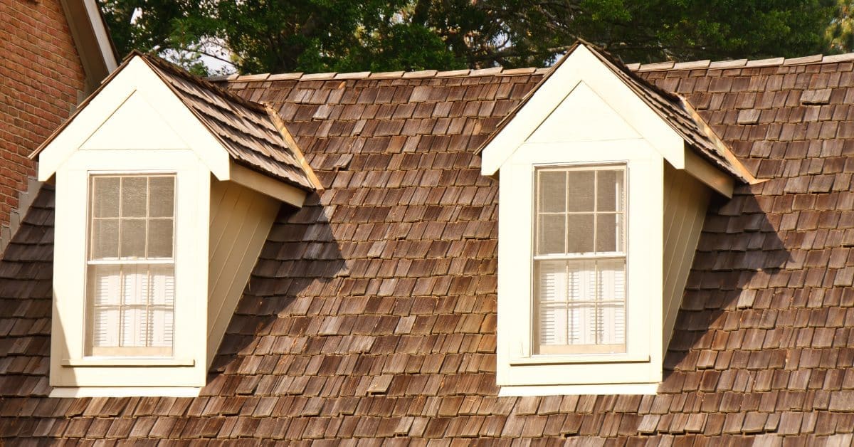 A house has rows of wooden shingles on its roof. Two windows with shades are visible as the sun shines brightly.