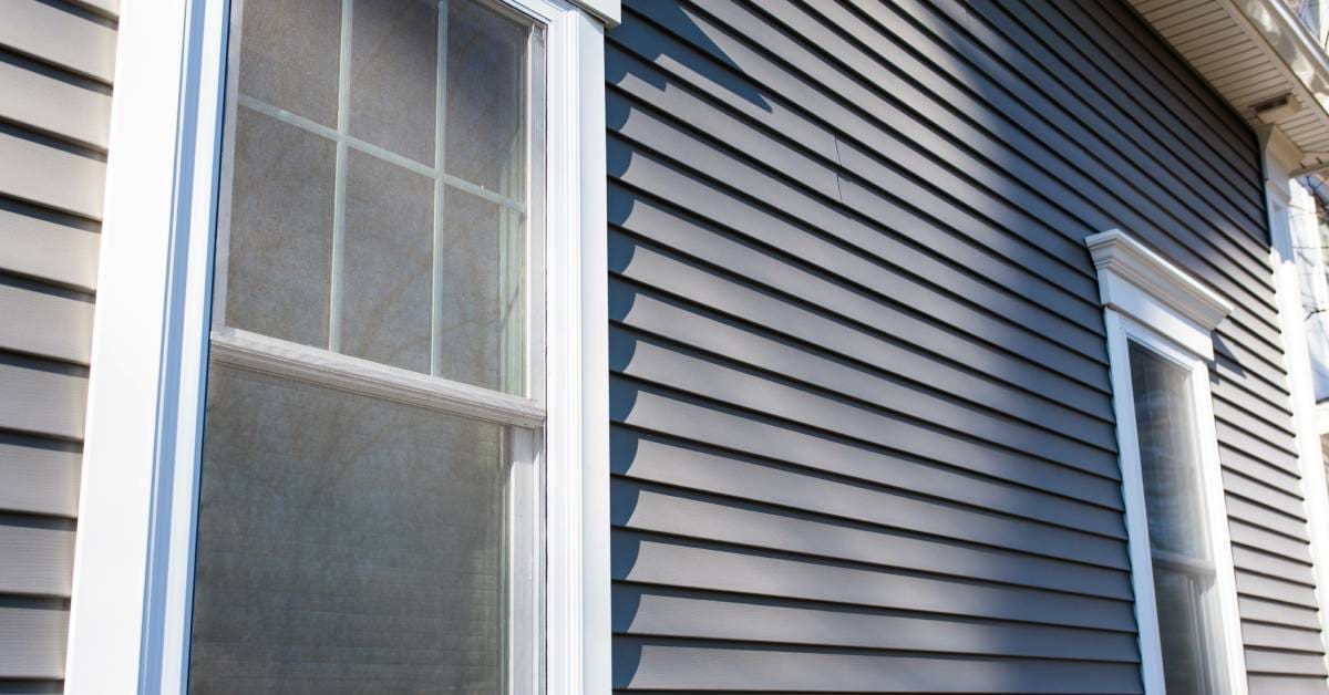 The side of a house with tall windows surrounded by white borders. Sunlight and shadow stretch across the gray vinyl siding.