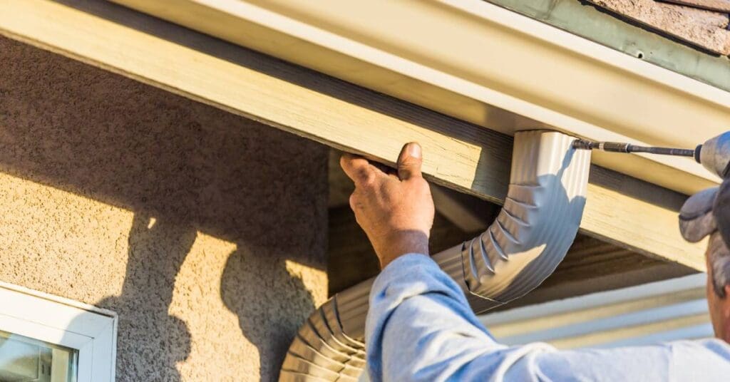 A man wearing a hat and a long-sleeve sweater is using a drill to attach a downspout to a brightly colored gutter.