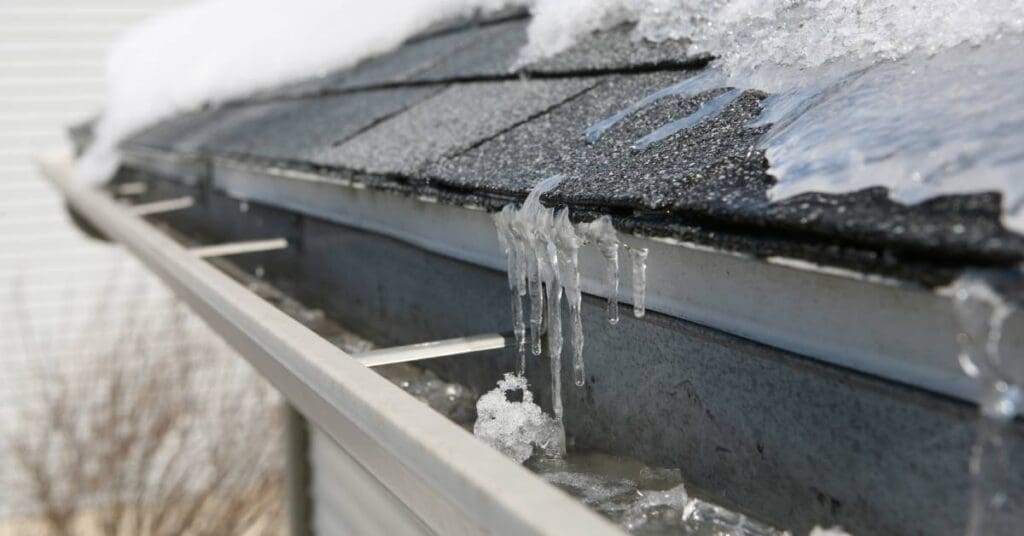 Snow and ice cover the black tiles of a roof's edge. Small icicles hang over the interior of a gutter system.