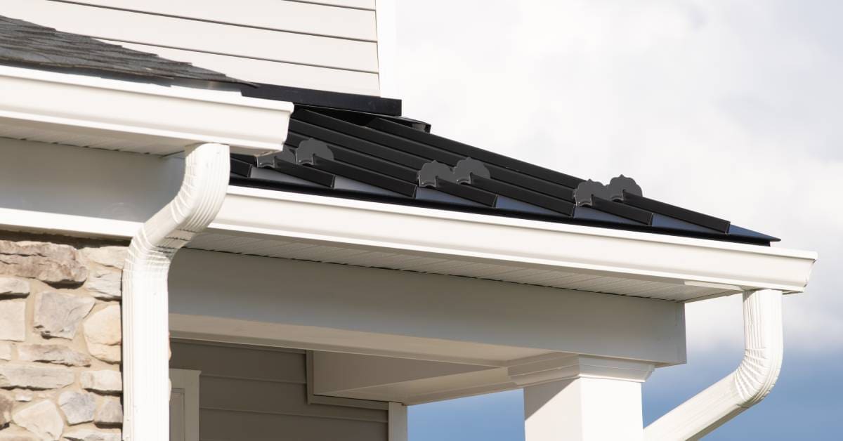 The corner of a house with black asphalt shingles and a white gutter system. A cloudy, blue sky is visible in the background.