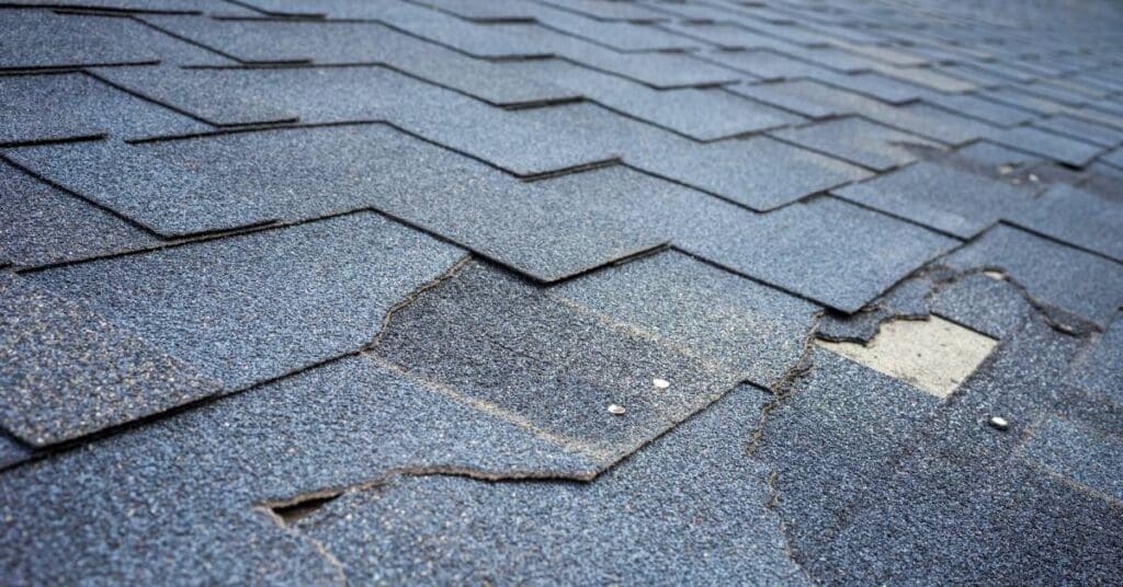 The gray asphalt shingles of a roof. Some shingles show signs of damage with chips and openings in the roof's surface.