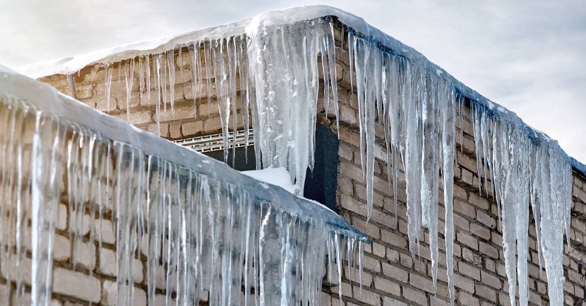 A house made of pale bricks has a thick ice dam and numerous large icicles hanging from the edge of the roof.