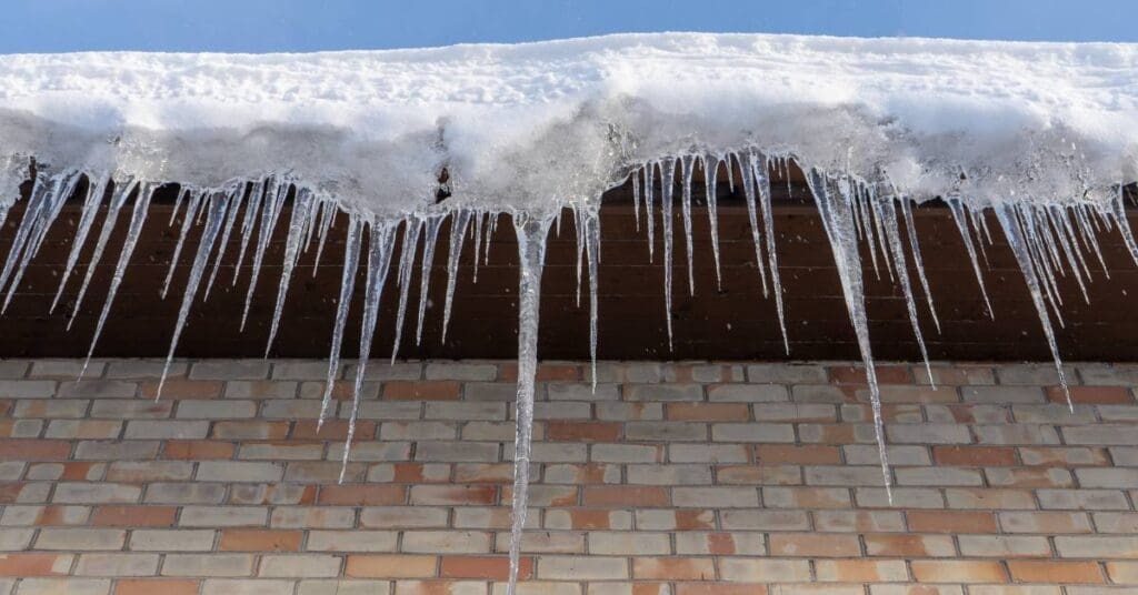 The edge of a brick building's roof is covered with a thick ice dam. Long icicles hang from the edge.