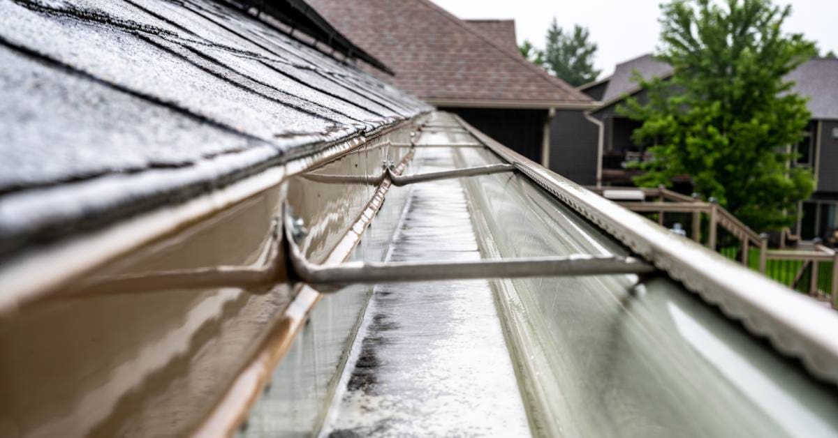 A roof gutter with water flowing throughout its metal drainage system with other residential homes in the background.
