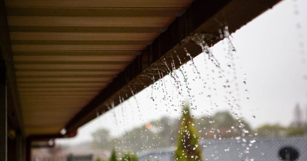 The underside of a brown roof's drainage system with multiple streams of water pouring along the length of the gutter.