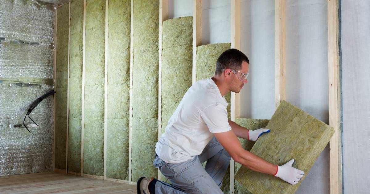 A man in a white shirt, gray pants, white gloves, and work shoes installing insulation into the wooden frame of a wall.