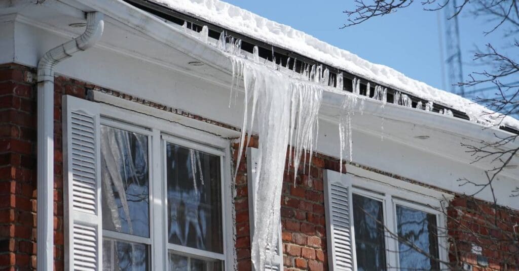 A brick house with a snow-covered roof showing signs of gutter damage as a long ice dam drags it down.