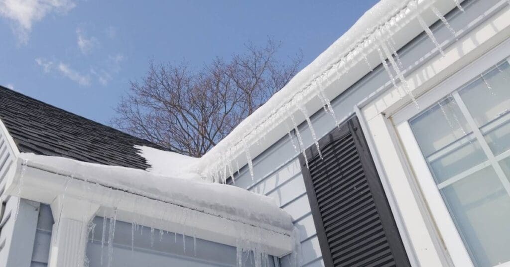 A house with black asphalt shingles and a large buildup of snow creating an ice dam and icicles along its edges.