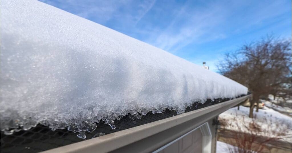 A roof covered with snow leading down to its edge stopping short at the gray gutters covered by a gutter guard.