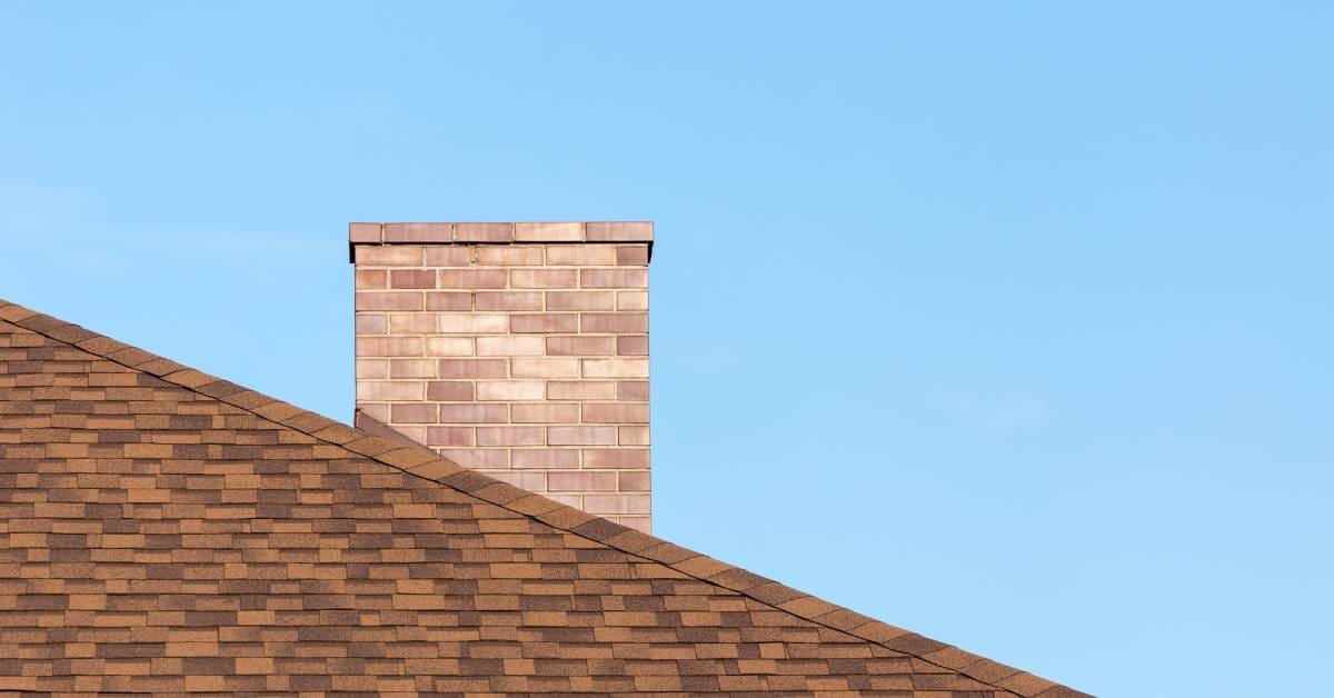 A roof with light brown asphalt shingles and a chimney made of pale brown bricks are illuminated by the sun.