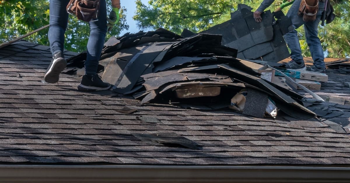 Two men are standing on a residential roof and removing damaged asphalt shingles while forming a pile.