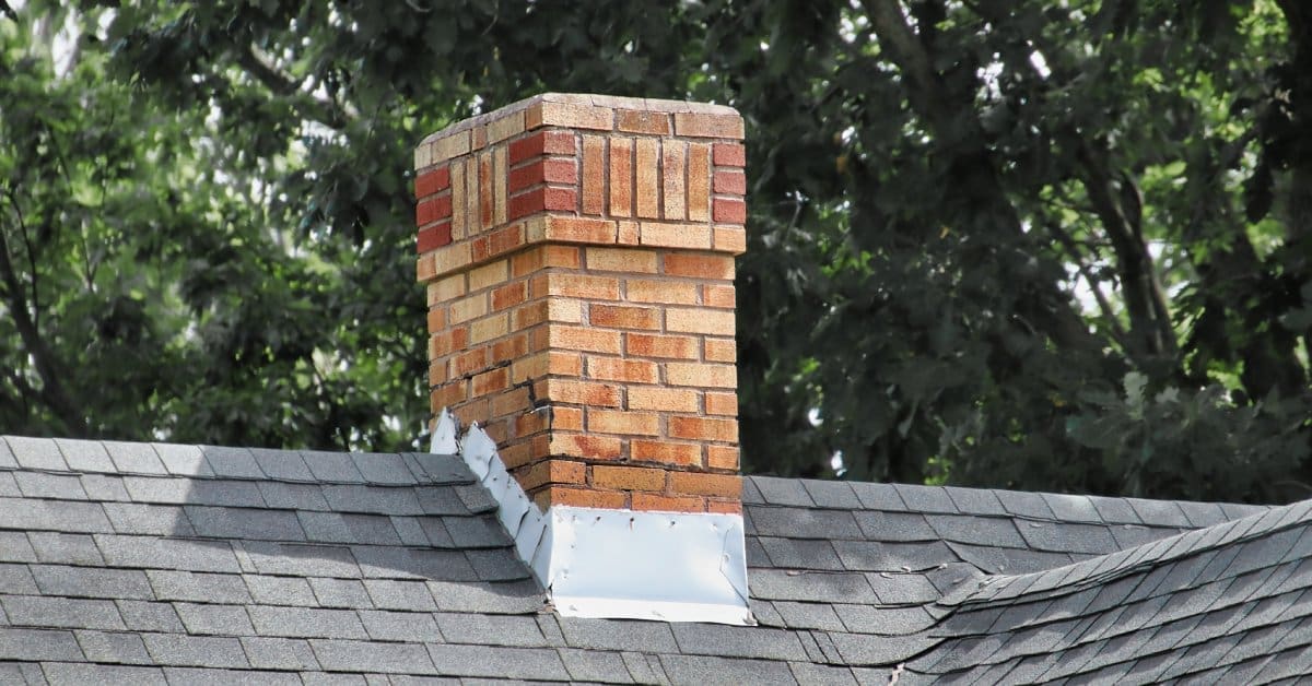A brick chimney with metal patches at its base sits on a roof with gray shingles. Trees are visible in the background.