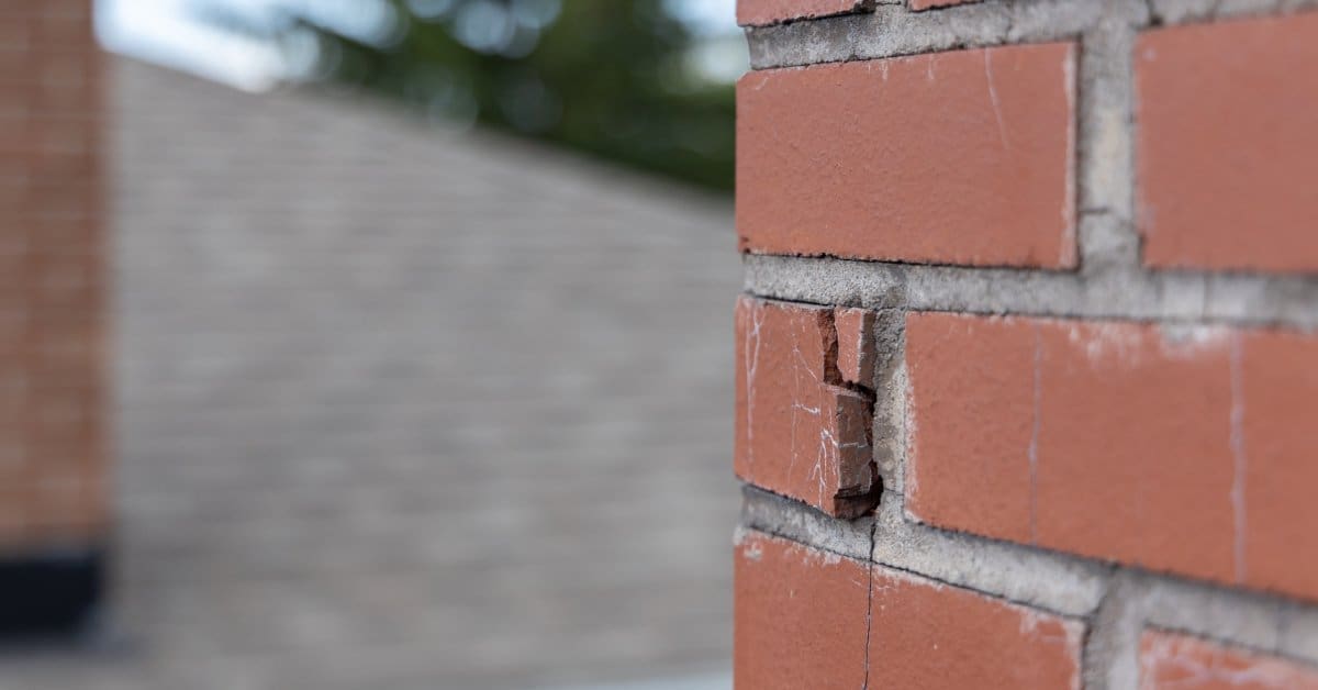A close-up of a red brick chimney with a visible crack. The roof of another house is in the background.