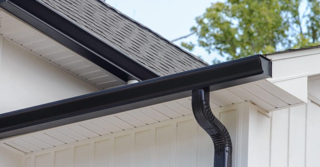 A black box gutter connects to a black downpipe with a curved bend. The gutter matches the black shingles on the roof.