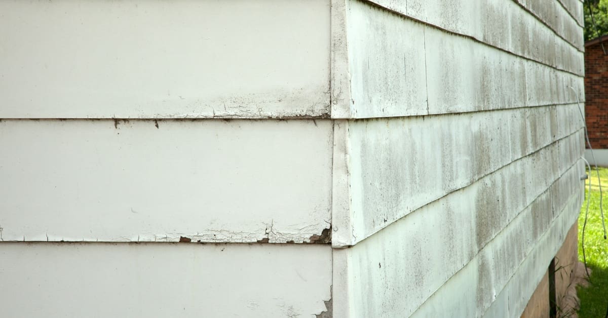 Cracked and peeling beige siding on the corner of a house, with dirt and grime accumulating along the surface.
