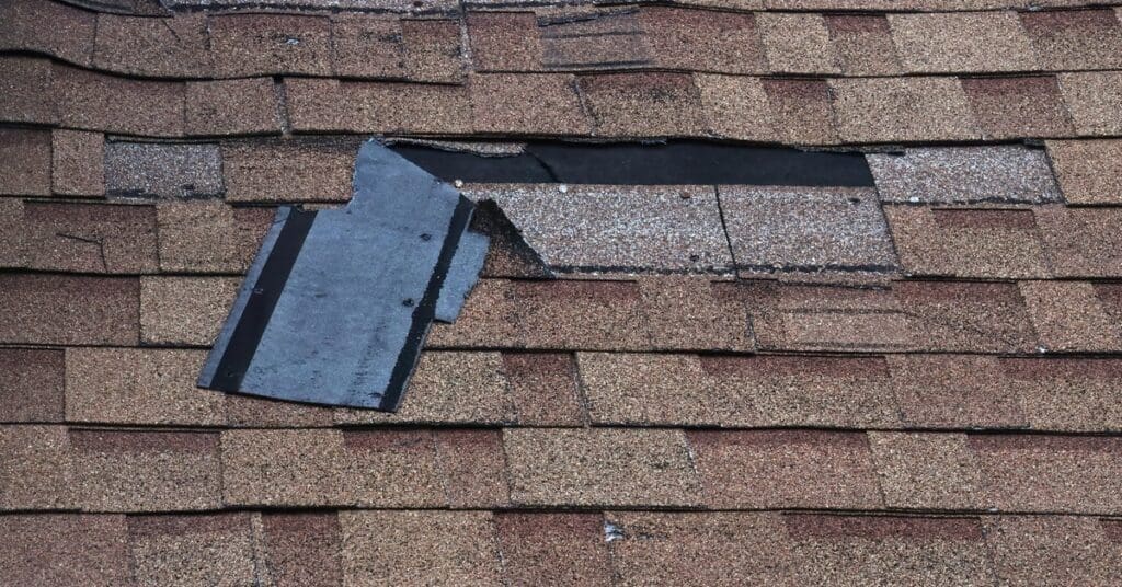 A roof with brown asphalt shingles. Multiple areas of the roof have shingles peeling off to reveal shingles underneath.