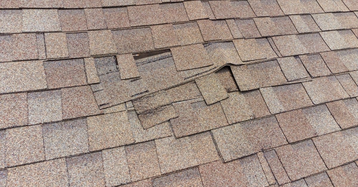 section of light brown asphalt shingles slips out of place on a roof, exposing a visible gap beneath.