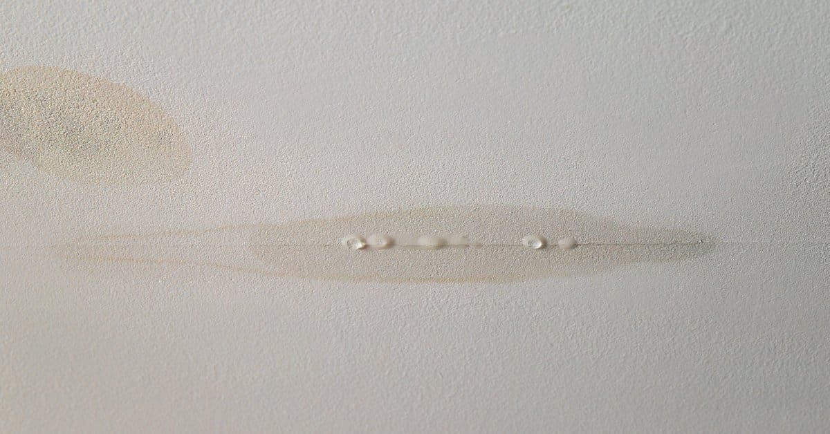 A small crack in a white ceiling shows droplets forming. Nearby, a round, light brown stain is visible.