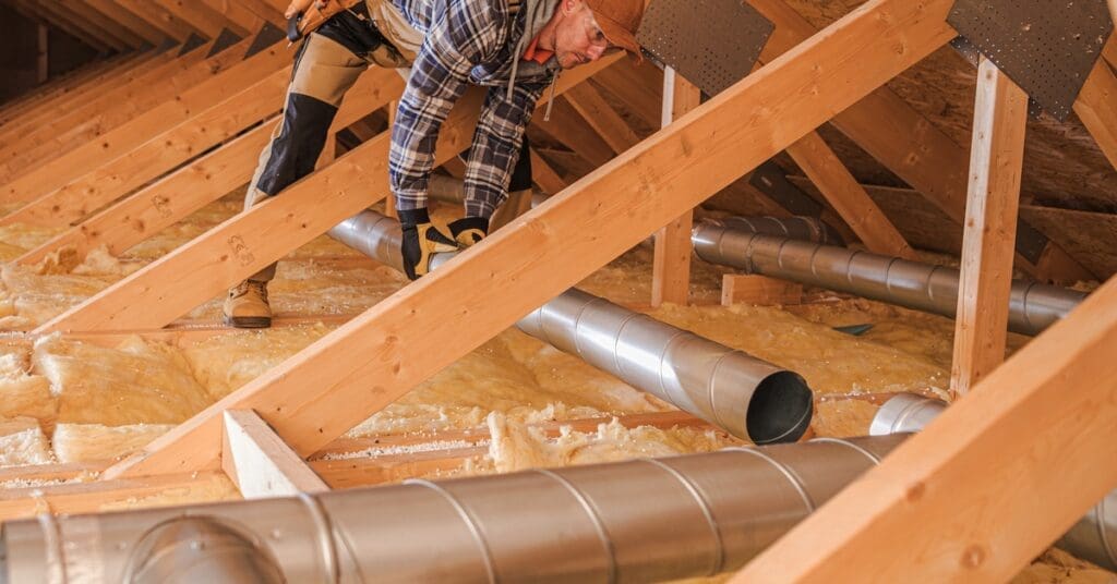 A man lays a section of air duct in an attic. Yellow insulation and wooden boards are visible beneath him.