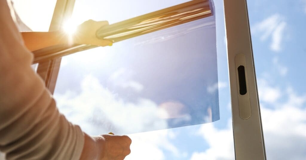 A pair of hands unrolls a sheet of dark film against a glass pane with sunlight and a clear blue sky in the background.