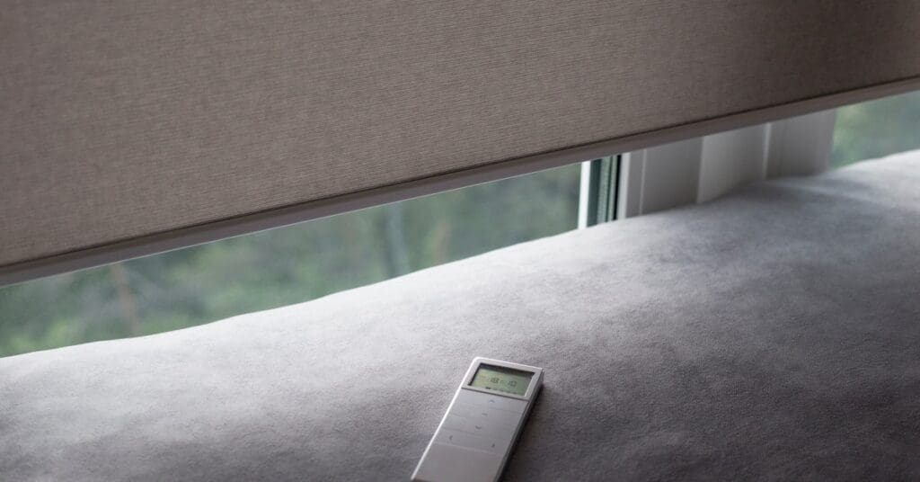 A gray fabric roller shade partially covers a set of windows, with a white remote control resting on a fabric surface.