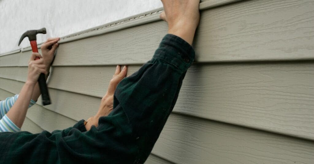 Two pairs of hands hold onto a strip of vinyl siding, with one pair holding it in place and the other about to hammer.