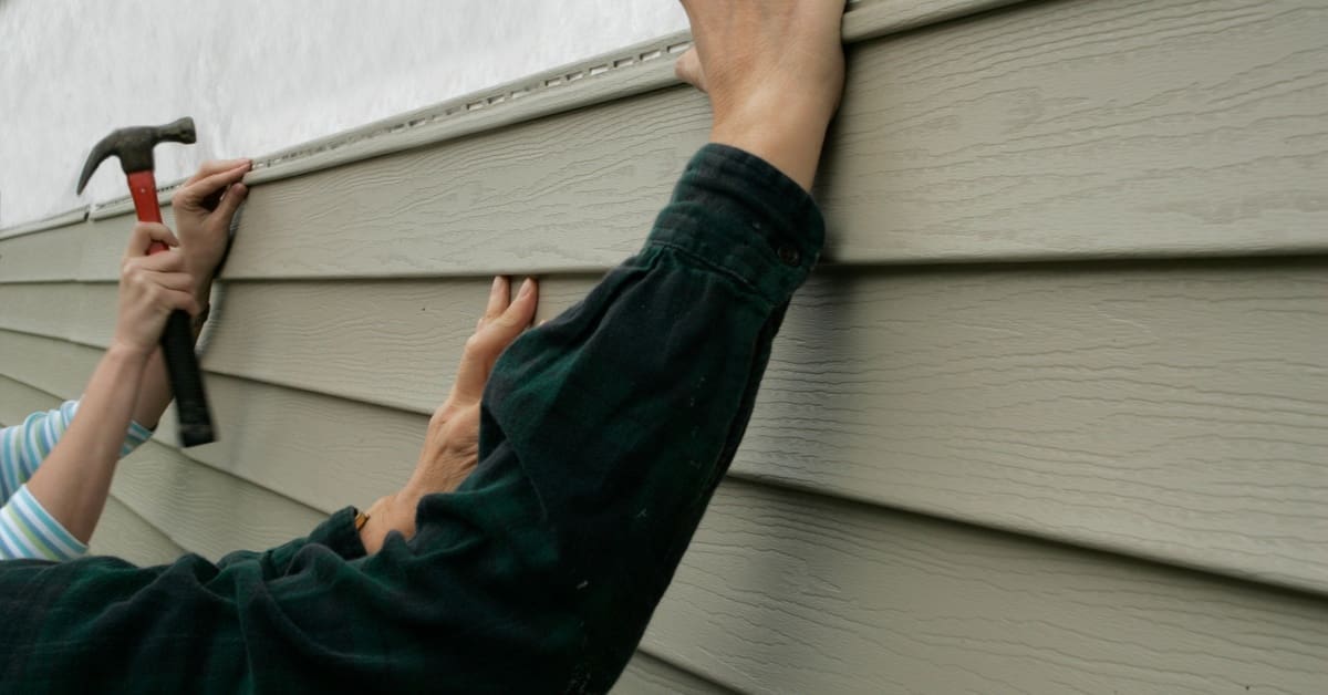Two pairs of hands hold onto a strip of vinyl siding, with one pair holding it in place and the other about to hammer.
