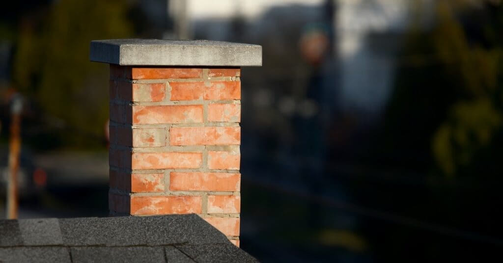 A brick chimney sits atop a roof with black shingles with blurred trees and a building in the background.