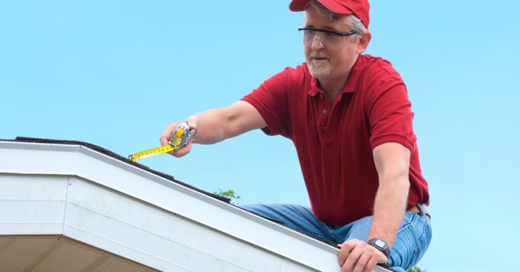 A man wearing a red shirt, matching cap, and safety goggles kneels on a roof while holding a measuring tape.