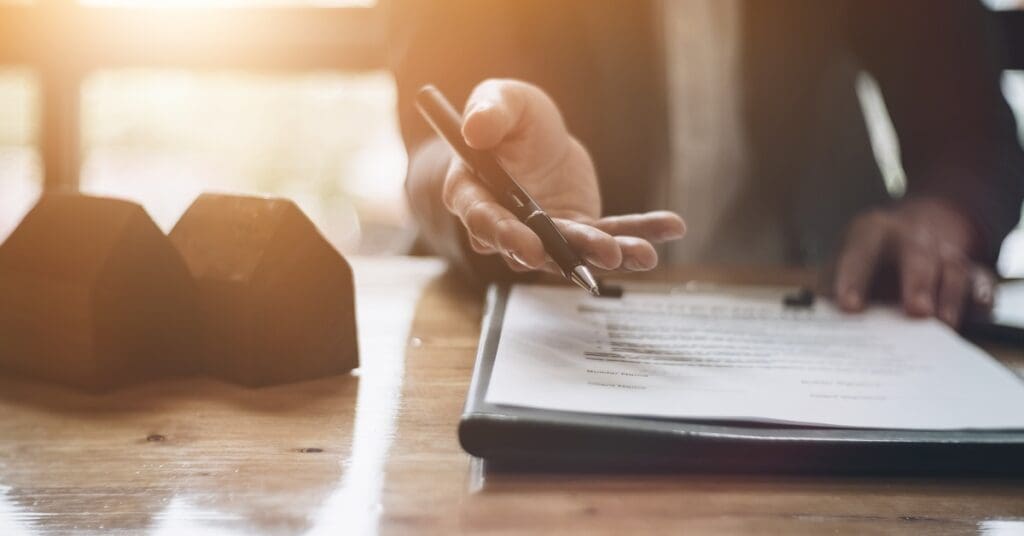A hand with a black pen hovers above a clipboard with paper beside two small model homes on a flat surface.