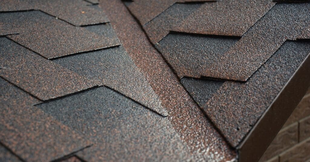 Brown valley flashing cuts between rows of matching shingles on a sloped residential roof with beige siding below.