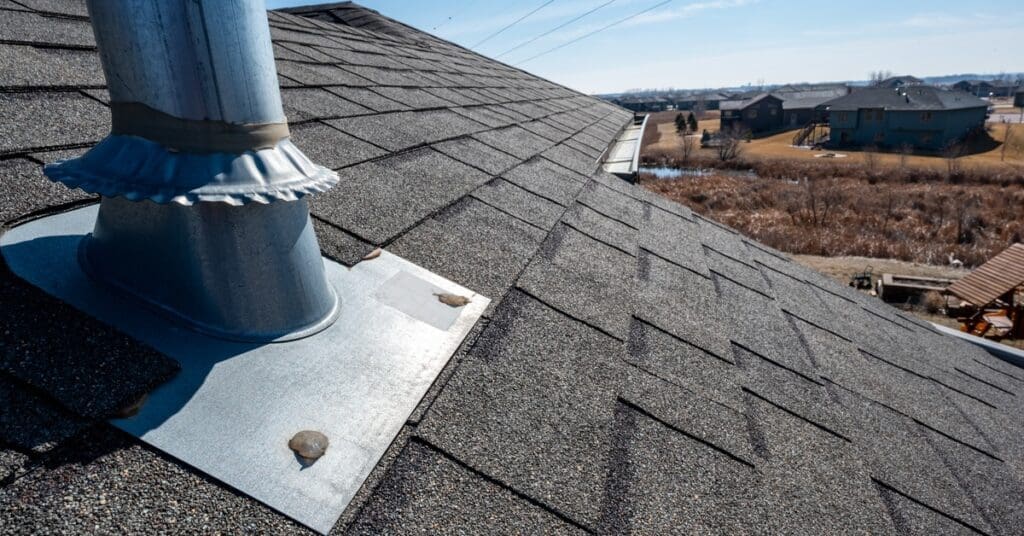 Metal flashing wraps around a roof vent pipe surrounded by black asphalt shingles under clear blue skies.