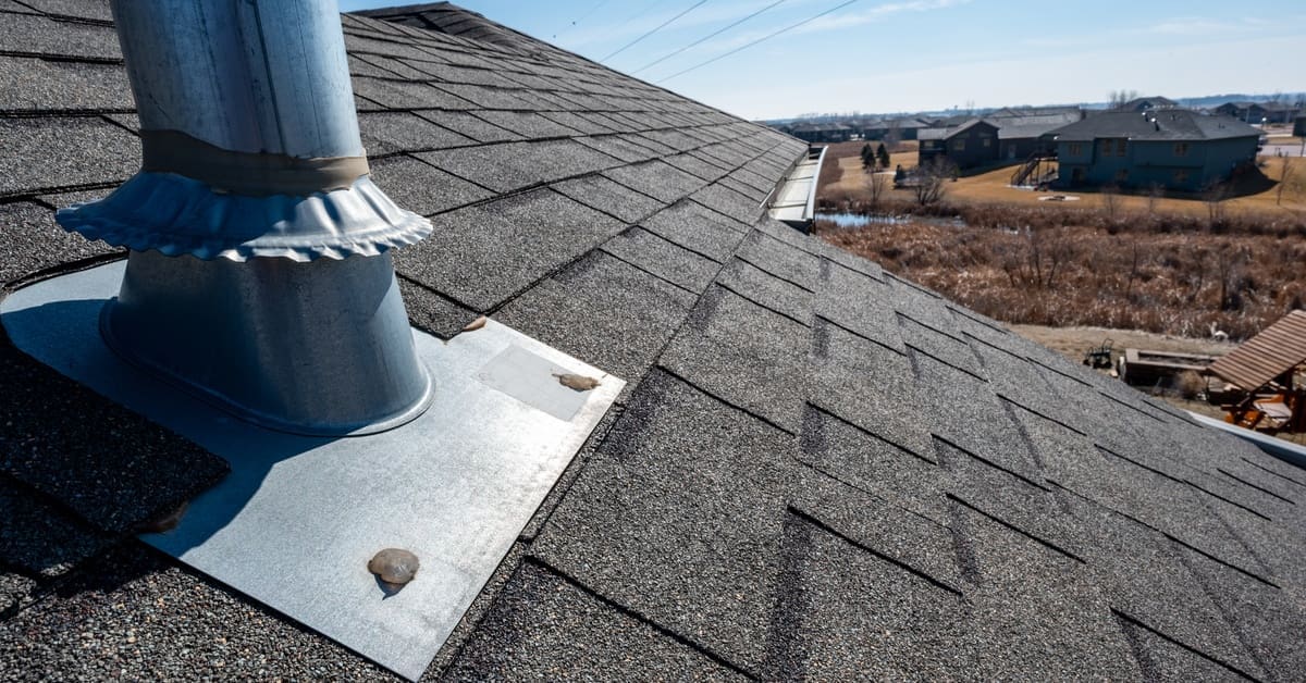 Metal flashing wraps around a roof vent pipe surrounded by black asphalt shingles under clear blue skies.