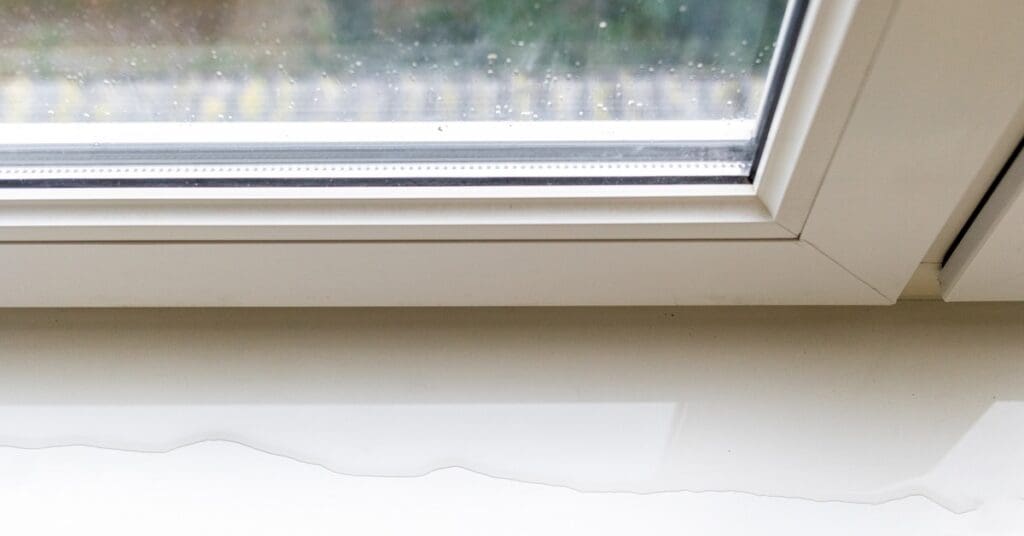 A close-up of the corner of a double-glazed window frame. There is a puddle of water on the white windowsill.
