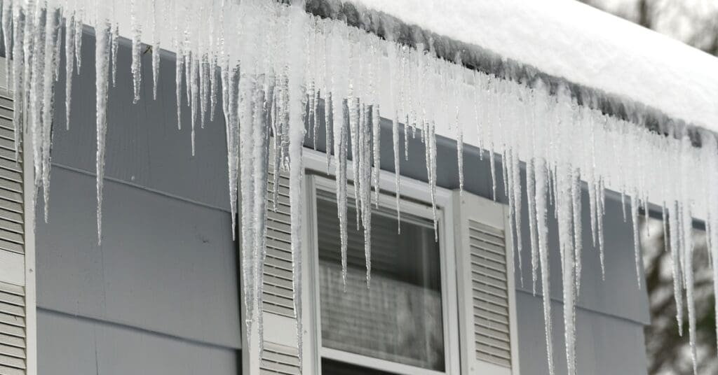 Large icicles hang from the roof edge of a blue house with white shutters and snow covering the roof.