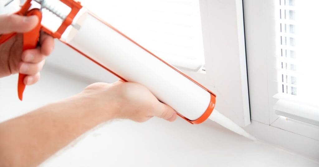 A person uses an orange caulking gun to seal the edge of a white window frame. The blinds on the window are open.