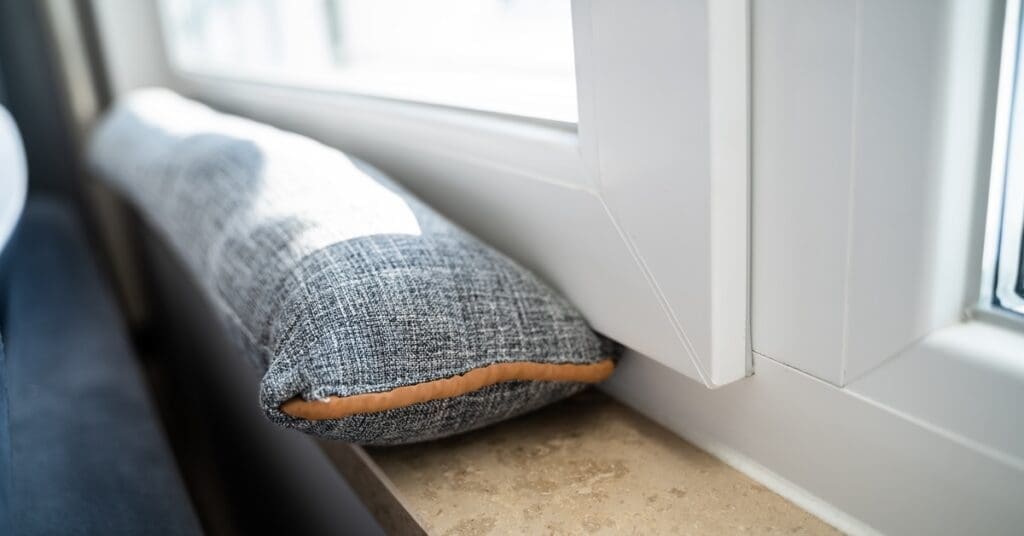 A gray draft excluder rests along a faux-wood windowsill above a blue couch. The window frame is white.