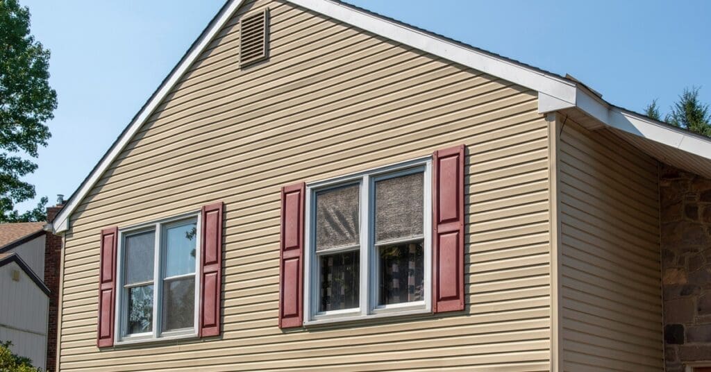 Sunlight reflects off a tan-sided house with red shutters and small indentations near a corner panel.