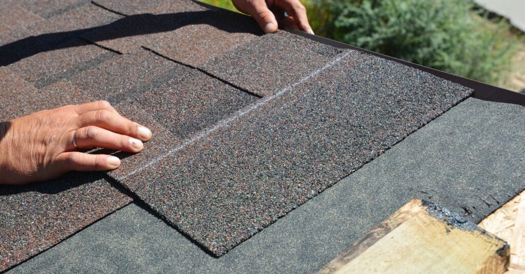 A person placing a reddish-brown asphalt shingle over faded black underlayment. Also nearby is a plank of wood.