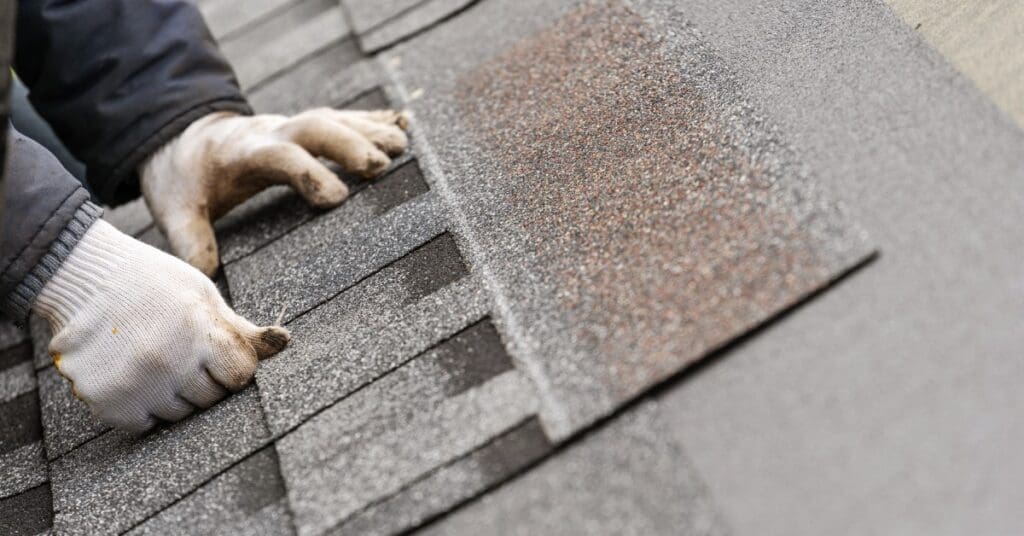 Gloved hands position an asphalt tile with black, gray, and white granules onto a roof's underlayment.
