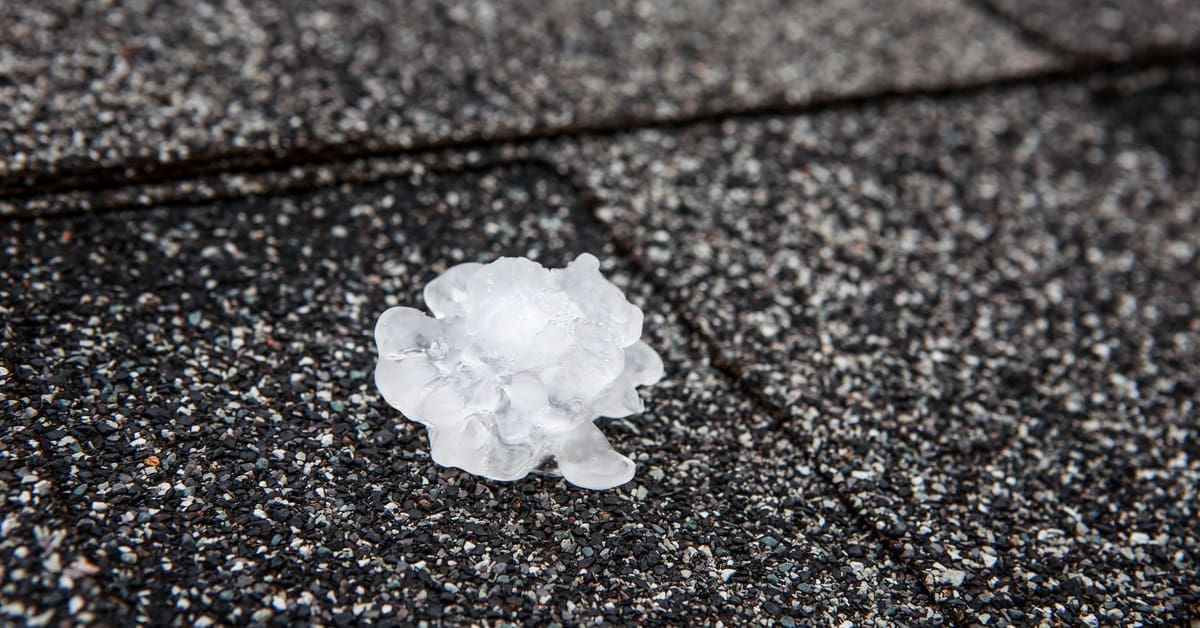 A chunk of hail sitting on black asphalt shingles speckled with white, gray, orange, and teal granules.