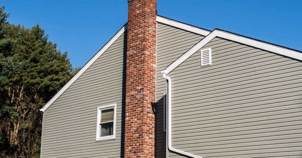 A gray house with a tall red brick chimney and a white downspout stands against a clear blue sky and trees.