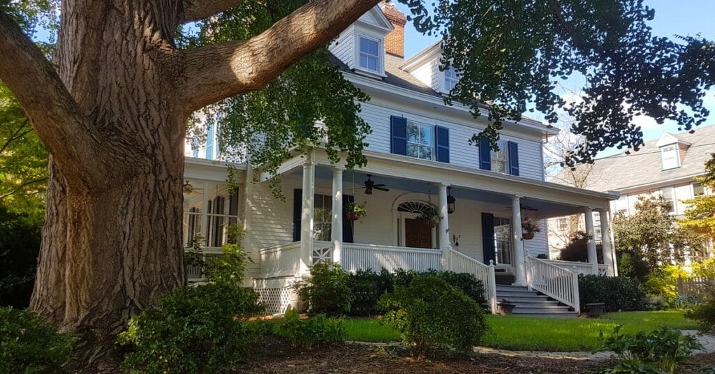 A large tree shades a white house with blue shutters, white railings, gray roof shingles, and a red brick chimney.