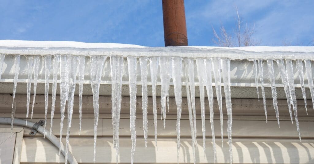Icicles hang over white box gutters on a beige-sided house with a red chimney vent and clear blue sky above.