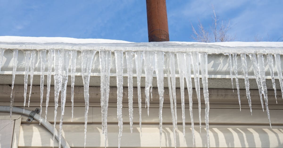 Icicles hang over white box gutters on a beige-sided house with a red chimney vent and clear blue sky above.