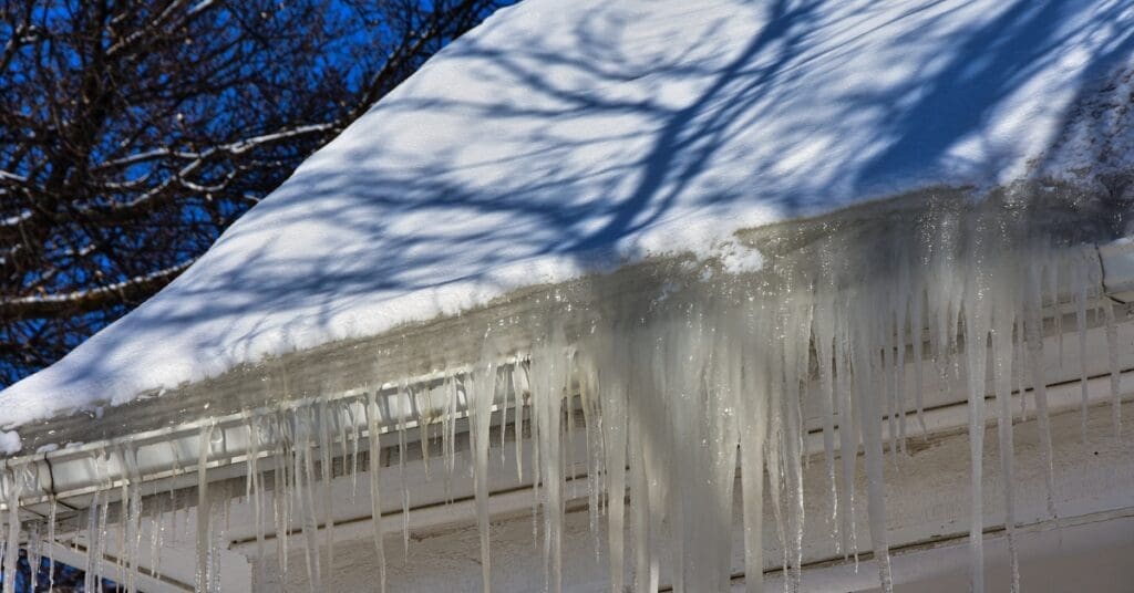 A sheet of snow covers a roof where thick ice dams have formed in the white box gutters of a residential home.